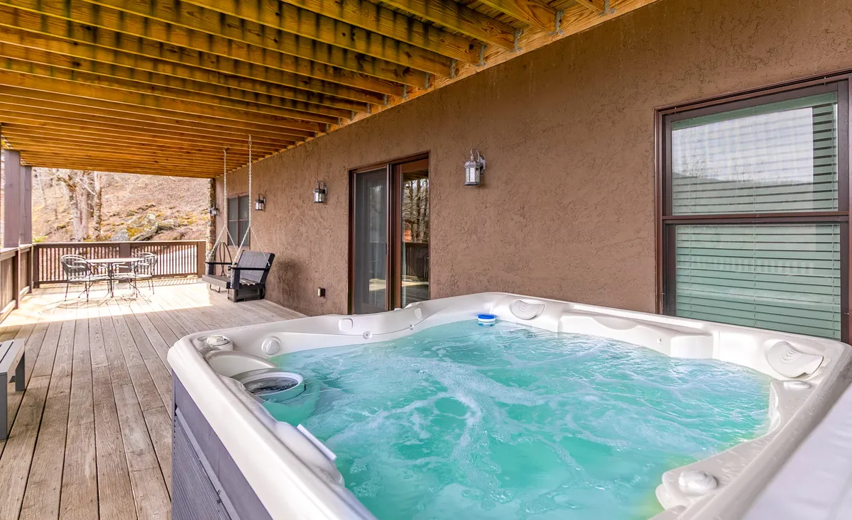 A spacious wooden deck with a prominent hot tub, surrounded by comfortable outdoor seating and a table set for meals. The scene captures a tranquil atmosphere with the sun setting in the background, emphasizing the relaxing and social nature of the space. The mountains can be glimpsed beyond the deck, reinforcing the connection to the Blue Ridge views.