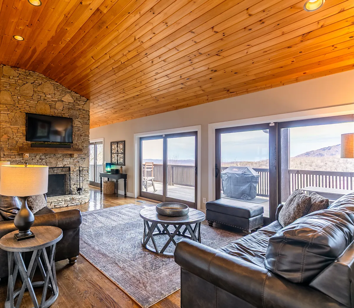 A spacious living area featuring a beautiful stone fireplace, large windows that allow abundant natural light, comfortable seating, and a warm wooden ceiling. The space feels both cozy and open, ideal for gatherings or quiet relaxation while enjoying panoramic views.