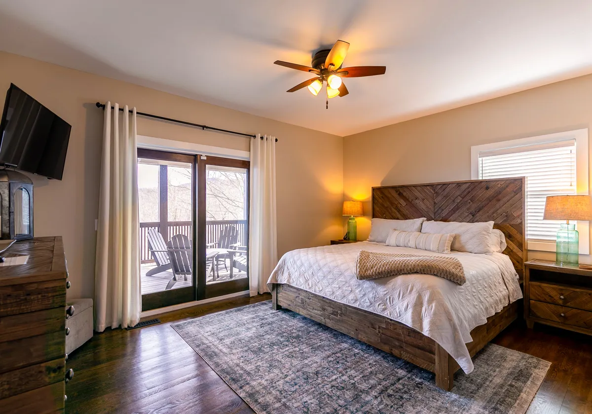 A spacious bedroom featuring a large, inviting bed adorned with decorative pillows, generous windows opening to a private balcony, a ceiling fan, a wooden dresser, and a soft area rug. Natural light fills the room, and the balcony offers the promise of tranquil outdoor moments.