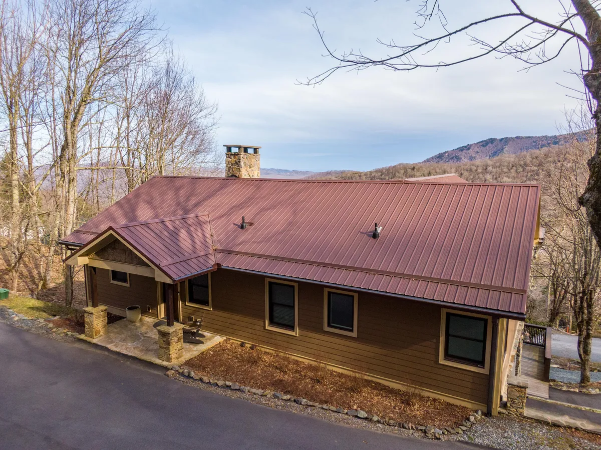 A broad exterior view of the house, featuring charming wooden siding, a sturdy metal roof, stone chimney, and tall trees framing the property. The porch invites guests to relax, and the surrounding landscape hints at space for outdoor play and gatherings.
