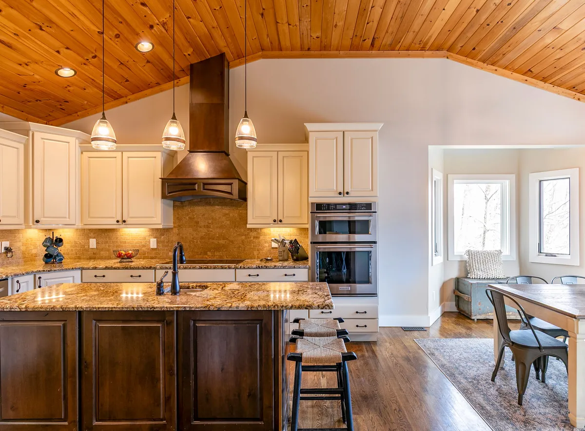 This image captures a beautifully designed kitchen with a granite island, modern stainless steel appliances, a wooden ceiling, and large windows letting in natural light. The layout invites gatherings and culinary experiences, emphasizing the luxurious yet welcoming ambiance of the home.