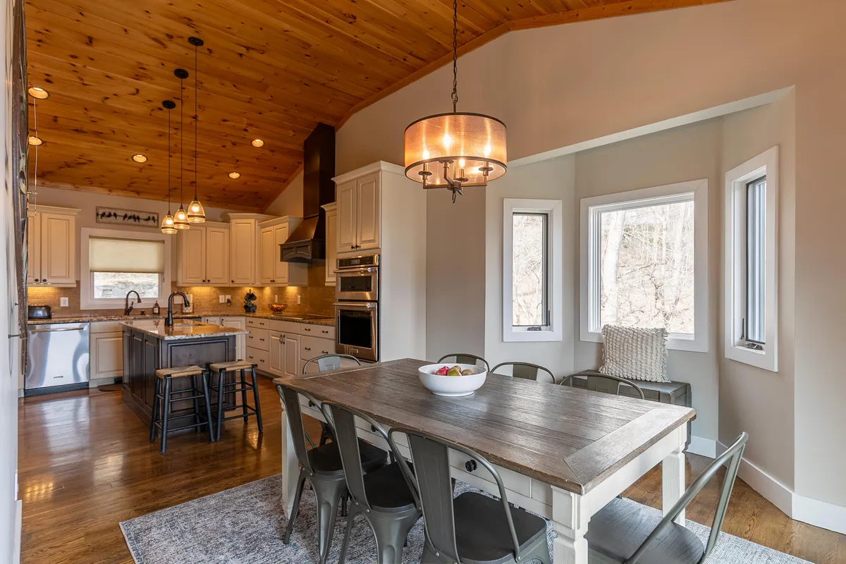 An open-concept space that seamlessly blends the dining area and kitchen. The dining table is positioned by large windows offering scenic views, while the kitchen island and wooden ceiling create a warm, welcoming environment perfect for meals and entertaining.