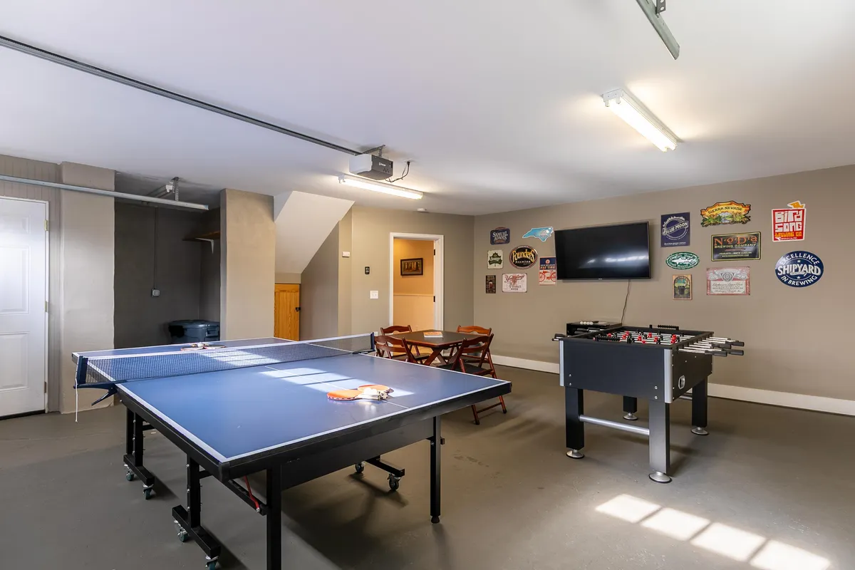A vibrant game room with a ping pong table and foosball table as focal points. A flat-screen TV is mounted on the wall, and decorative signs add a playful, welcoming touch to the space. The arrangement is open, inviting guests to gather for friendly competitions and socializing.
